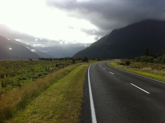 new zealand road, cloudy day, grass fields beside road, mountain range in the distance.