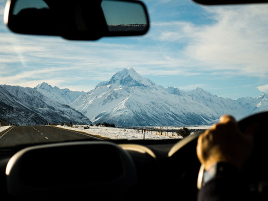 blue skies with clouds, person driving car towards mount cook nz, snow capped mountain range in view.