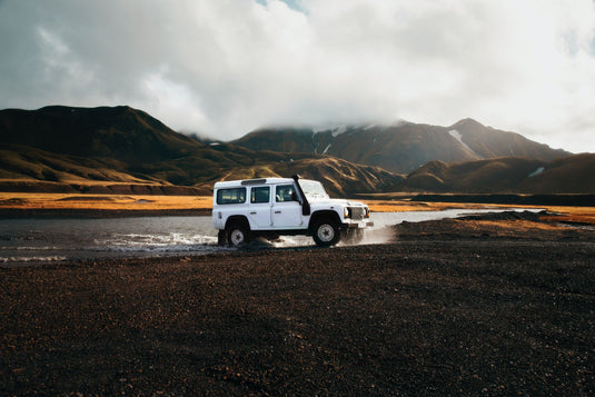 land rover defender driving out of river, mountains in the background, cloudy day.