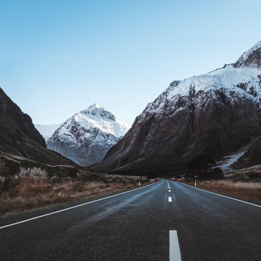 low angle of road heading into distance, beside snowy rugged mountains, blue skies.  