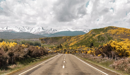 road driving through nz busy, snow capped mountains in distance, clouds 