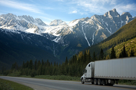 long haul truck driving on forrest lined open road, rugged snowy mountains and blue skies