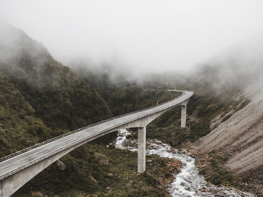 road bridge weaving through a valley, river running below and gravelly mountain wraps around, misty cloud covering.